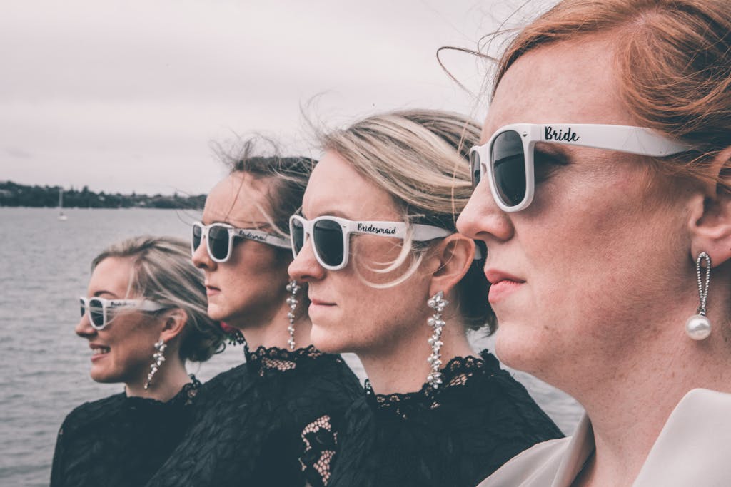 Side view of crop creative bride with red hair in elegant dress and sunglasses standing with happy bridesmaids on seashore against cloudy sky before wedding ceremony