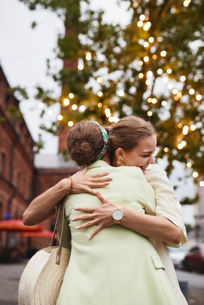 Two adult women joyfully hug outdoors with lights in the background, conveying friendship and happiness.